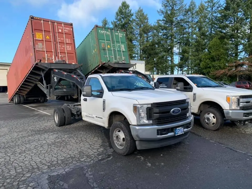 Two white trucks with shipping containers tilted on their beds, ready to slide off